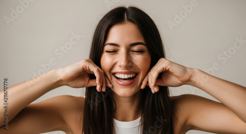 Portrait of joyful young brunette woman laughing with closed eyes while touching her cheeks against plain studio background for dental health.