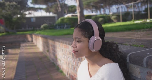 Teen sitting on brick wall at park, listening via pink headphones, mouthing to music, copy space