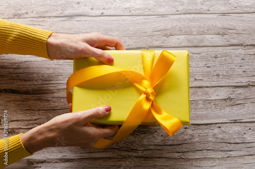 Woman's hands holding a yellow gift box with yellow ribbon on a wooden table.