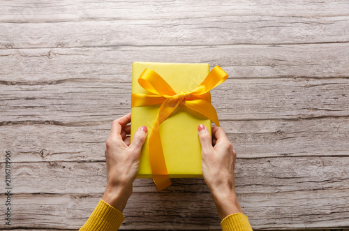 Woman's hands holding a yellow gift box with yellow ribbon on wooden table with copy space.