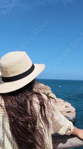 Young woman with long hair wearing a straw hat while enjoying a sunny day on a cliffside, gazing at the beautiful ocean and rocky shoreline from a viewpoint on her summer vacation