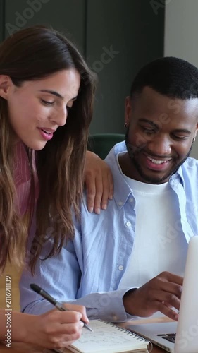 Smiling young multiracial couple working from home, with the man typing on a laptop and the woman taking notes in a notebook while they discuss their project and collaborate as a team