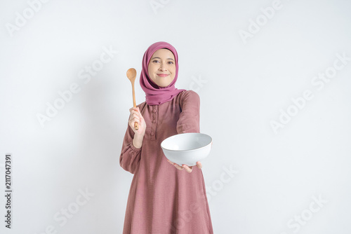 Fotografía Smiling Asian Muslim woman wearing hijab holding empty white bowl and wooden spoon offering food isolated on white background
