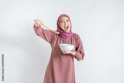 Cuadro en lienzo Cheerful Asian Muslim woman raising wooden spoon with excited mouth open holding white bowl isolated on white background