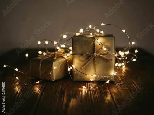 Two small gift boxes wrapped in gold paper and tied with twine, illuminated by warm glowing fairy lights on a dark rustic wooden table, creating a cozy and intimate atmosphere for holiday celebrations