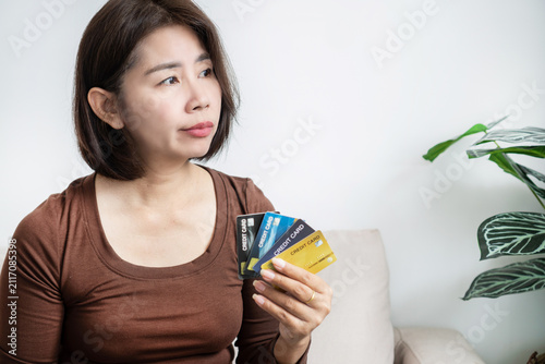 Canvastavla Worried Asian woman holding multiple credit cards with hesitant expression at home