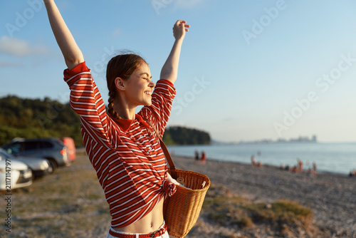 Fotografie Young woman on a sunny beach raises her arms in joy while holding a woven basket, wearing a red striped shirt tied at the waist, basking in warm casual summer mood