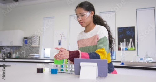 Schoolgirl putting on goggles checking tablet at lab placing green cup in rack, examining liquids