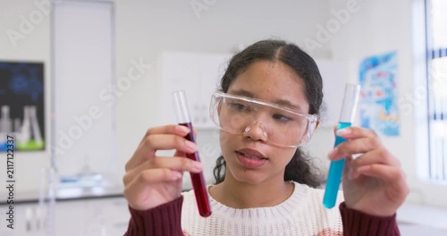Teen student wearing goggles comparing red and blue test tubes in lab after teacher cue, explaining