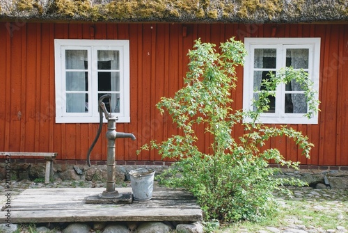 Red Scandinavian Cottage With Vintage Water Pump and Green Shrub Outside White Windows