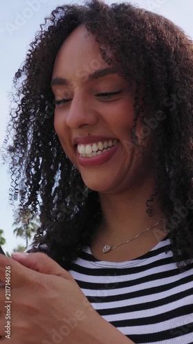 Vertical HD video of young Latin American girl using mobile phone outdoors. Happy young Brazilian woman checking smartphone in a sunny day. Technology and lifestyle concept. 