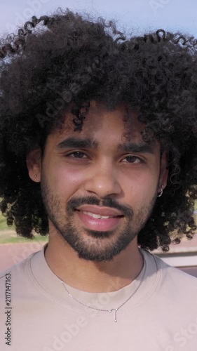 Portrait of smiling young Latin American man looking at camera outdoors. Confident African American millennial with curly hair enjoying a sunny day in the city. Vertical HD video frontal headshot.
