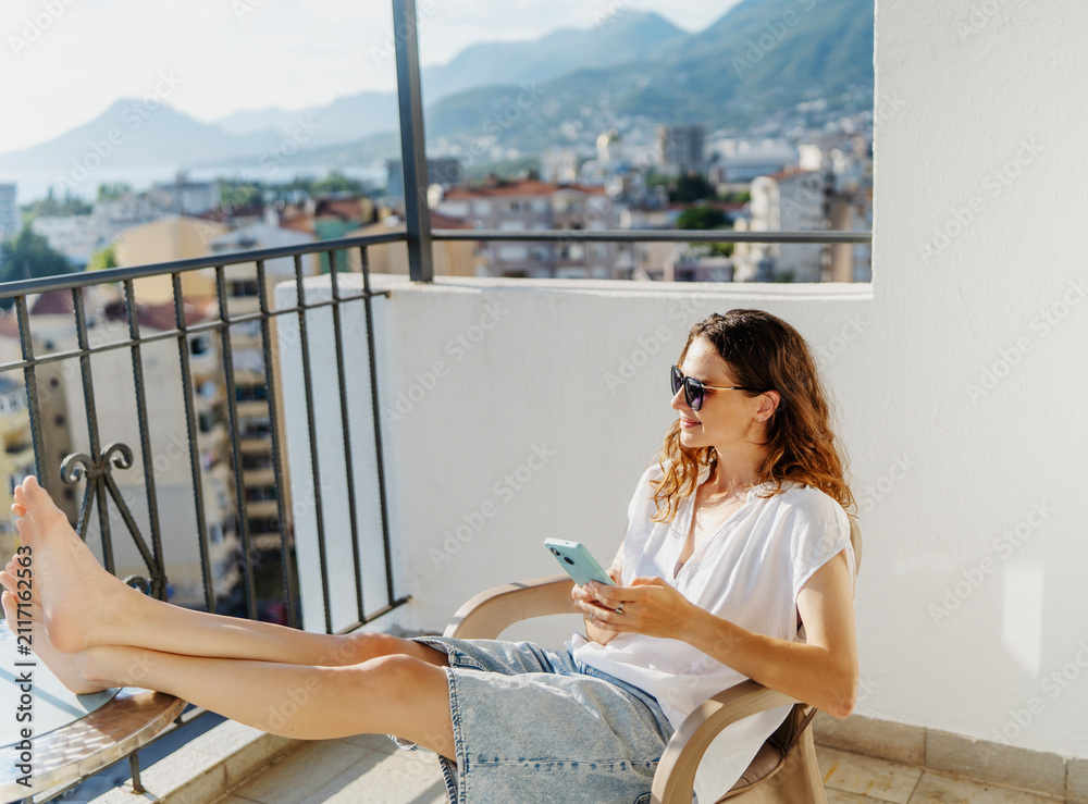 Obraz premium Happy young woman sitting on balcony chair with barefoot legs on table and looking at smartphone screen.