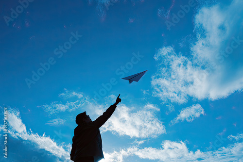 Silhouette of passionate young man throws folded paper airplane toward beautifully bright cloudy blue sky, expressing deep desire for total freedom, creative startup ideas, and genuine hope