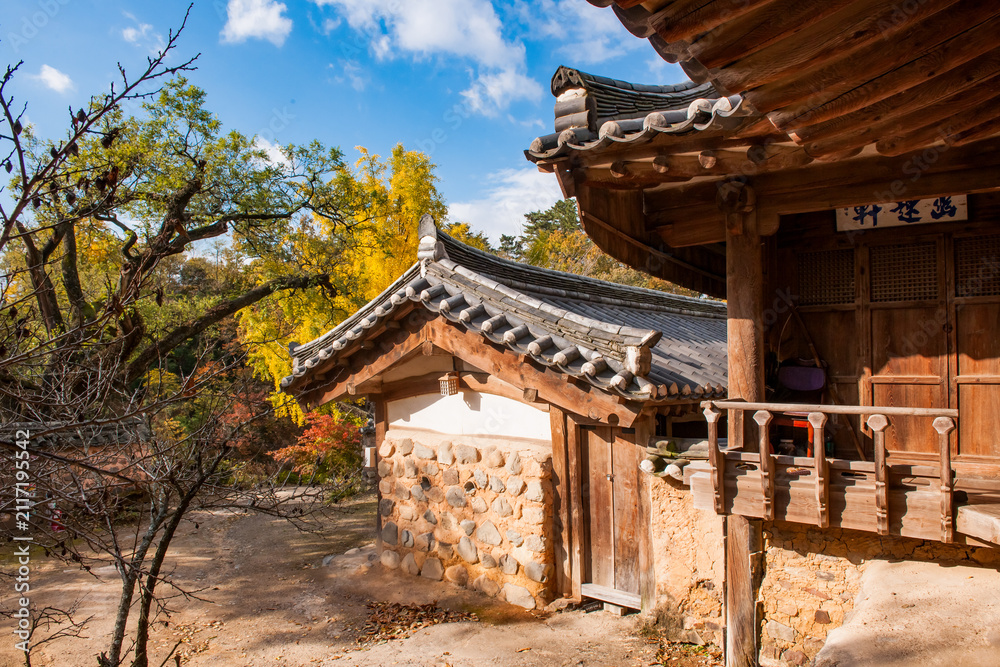 Fototapeta premium Scenic Exterior View of Traditional Korean Hanok Architecture Featuring Tiled Roofs, Earthen Wall, and Vibrant Autumn Foliage Under Blue Sky