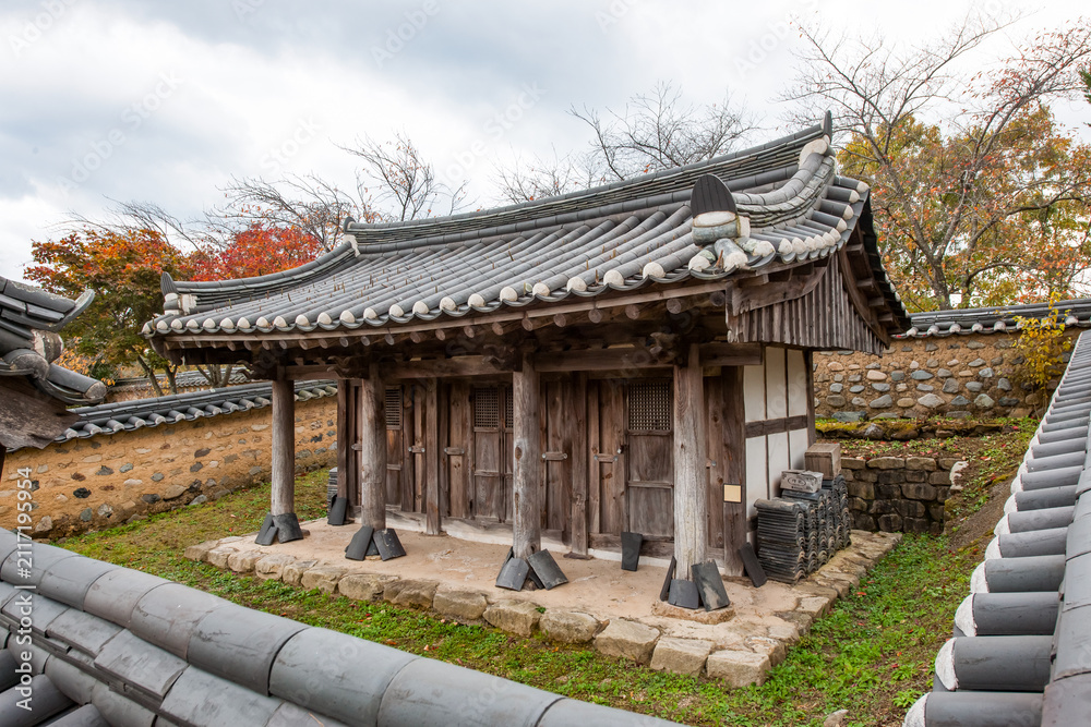 Fototapeta premium Exterior View of Traditional Korean Hanok Building Featuring Wooden Porch, Curved Tiled Roof, and Foreground Garden Plants Under Sky with Clouds