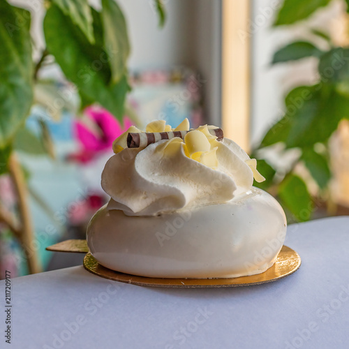 Elaborate single-serve cake with white frosting and chocolate decoration sits on a golden base. Plant bokeh background.