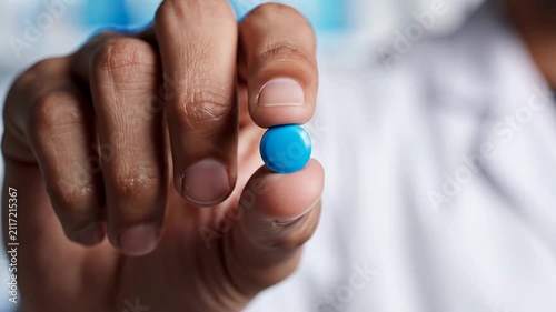 A professional doctor wearing a white medical gown carefully holds one small blue medicinal tablet between fingers to demonstrate the effective pharmaceutical treatment for patients battling