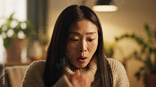 A distressed East Asian woman wearing a cozy high neck knit sweater experiences a sudden medical emergency while struggling for air inside her warm home during a dark and quiet winter evening