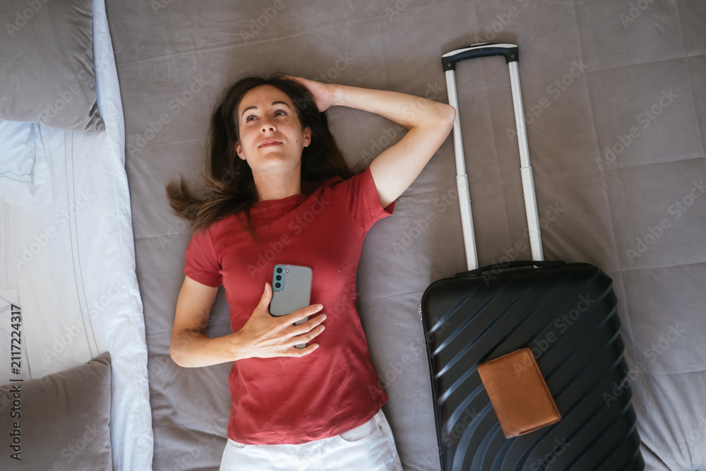 Fototapeta premium Woman lying on a hotel bed with a smartphone, suitcase, and passport holder, looking up thoughtfully, dreaming about upcoming vacation or business trip adventures