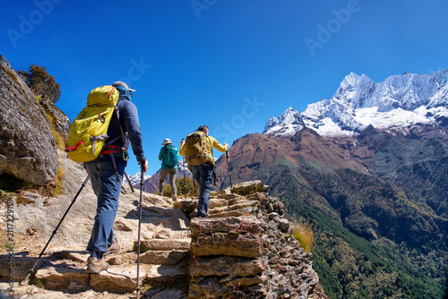 Group of hikers with backpacks and poles climbing ancient stone steps on trail to Everest Base Camp in Nepal, with snow-capped mountain peak.