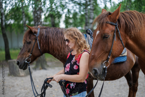A woman with sunglasses stands holding the reins between two brown horses outdoors near trees