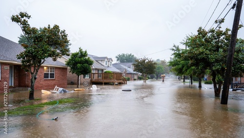 Flooded residential street after heavy rainfall with water covering road and front yards of brick houses in suburban neighborhood during daylight storm event.