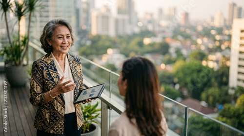 Two businesswomen converse on a balcony overlooking a cityscape, one holding a tablet.