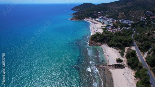 Aerial drone view of a crystal clear turquoise bay on Thassos Island, Greece, with swimmers, paddleboards and a sandy beach surrounded by rocky coastline on a sunny summer day.