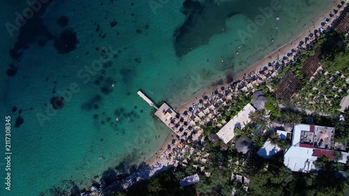 Aerial drone view of a crystal clear turquoise bay on Thassos Island, Greece, with swimmers, paddleboards and a sandy beach surrounded by rocky coastline on a sunny summer day.