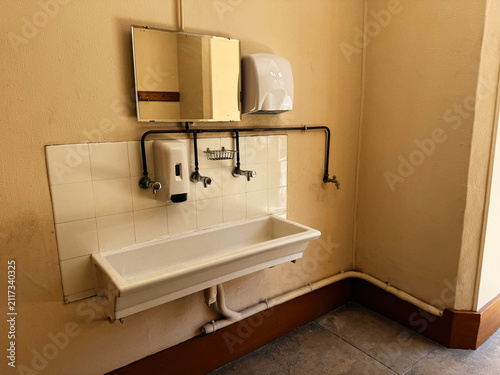 Vintage communal sink and soap dispensers in a tiled restroom corner in Paris, France with mirror, exposed pipes and warm plaster walls