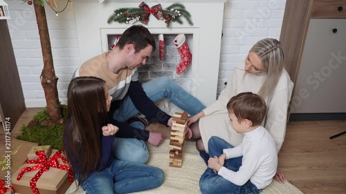 Happy family playing board game at Christmas, parents with kids stacking wooden blocks tower on floor near decorated fireplace, cozy winter holiday home activity, table game concept