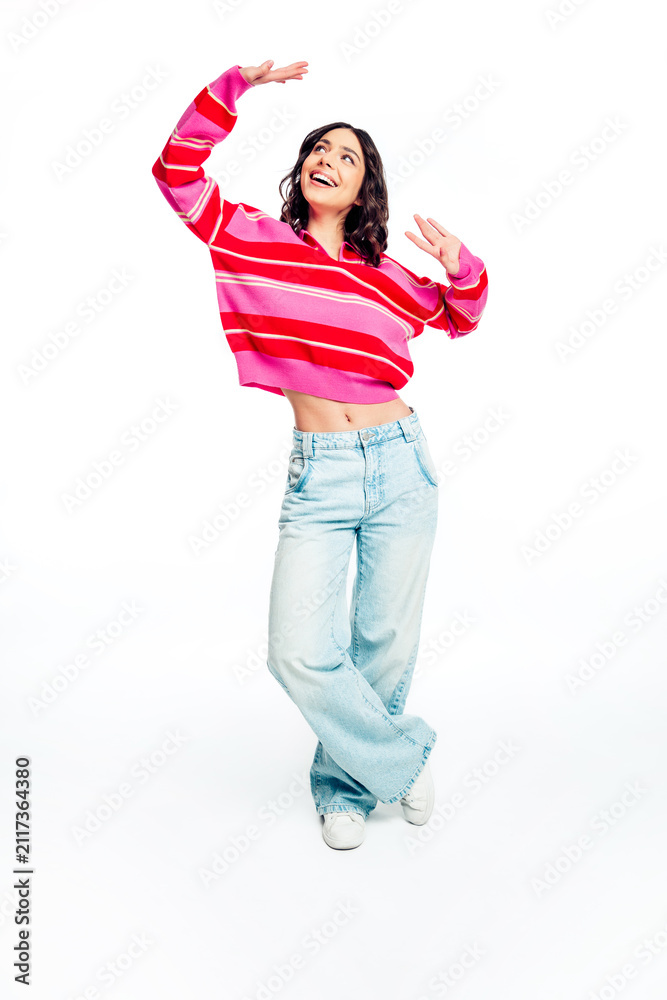 Obraz premium Happy young woman dancing in a pink striped sweater and wide‑leg jeans smiling against a white background