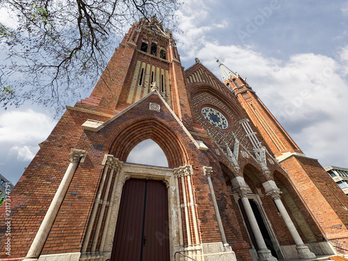 Imposing gothic revival style red brick cathedral featuring a towering facade, pointed arch entrance, intricate stone columns, and a large circular rose window.