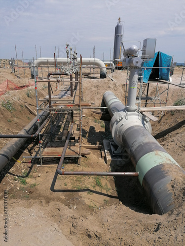 Industrial construction site featuring large gas pipelines laid inside a deep trench with metallic scaffolding, staging platforms, and heavy infrastructure equipment.