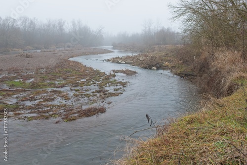 Winding river flows beneath high riverbank during foggy day