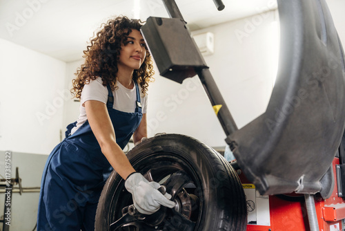 Cuadro en lienzo Female auto mechanic with curly hair wearing blue overalls works on a tire using