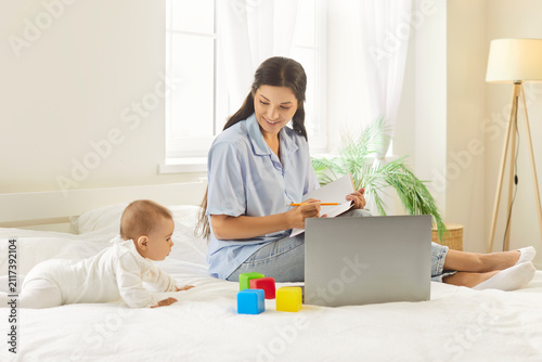 Fotografie Mother working on a laptop while her baby crawling among bright blocks on the bed, blending childcare and productivity at home in a cozy room