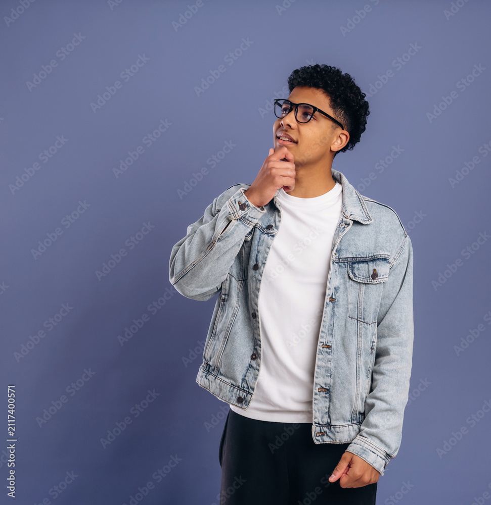 Fototapeta premium Young African American male wearing denim jacket and glasses poses thoughtfully against a solid purple background, showcasing a casual and modern style