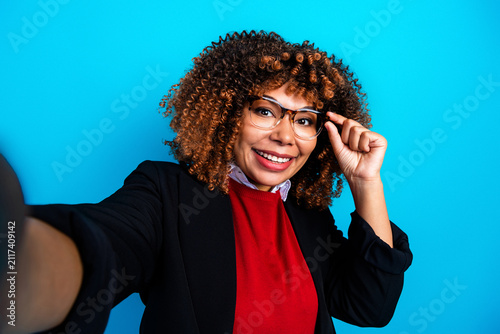 Fotografie Smiling young professional woman taking a selfie in glasses against a bright blu