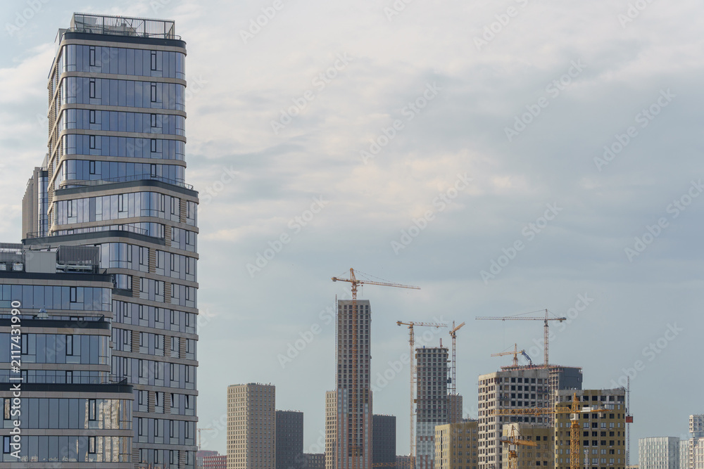 Fototapeta premium Construction site with tower cranes and unfinished high rise buildings next to a modern glass skyscraper. New residential district development area under cloudy sky.