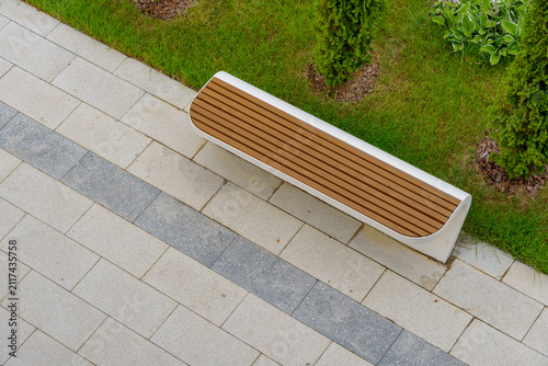 Wooden park bench on a stone tile pavement next to a green lawn with shrubs. Modern outdoor street furniture in a landscaped residential courtyard area from a high angle view.