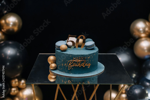 Blue birthday cake with golden decorations and number 50 on a mirrored table surrounded by black and gold balloons.