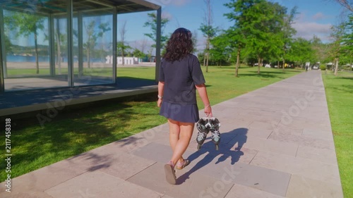 A woman enjoying a sunny day walks along a scenic path, holding roller skates in her hand, ready for an adventurous ride amidst lush greenery and water views.