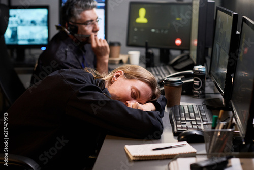 Fotografía Caucasian woman sleeping at control desk while male coworker monitoring screens, showing workplace fatigue and burnout