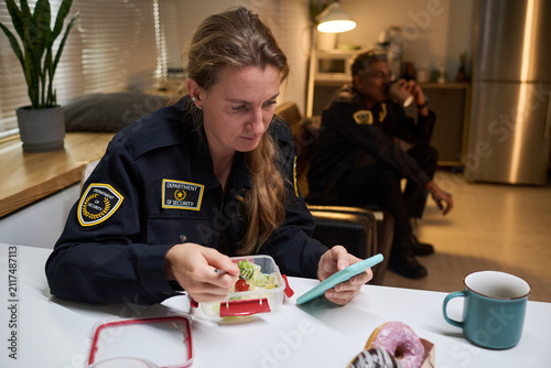 Caucasian security woman checking smartphone while eating meal during work break, colleague resting behind. Ideal for shift work, workplace fatigue, security staffing campaigns