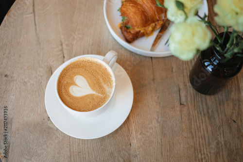 Overhead view of a white cup of cappuccino with delicate heart latte art, with a blurry plate containing a croissant sandwich and a dark vase with flowers in the background.