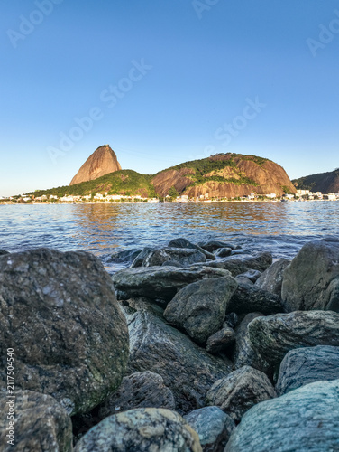 Aerial view of Sugarloaf Mountain rising above the rocky shoreline and calm waters in Rio de Janeiro, Rio de Janeiro, Brazil.