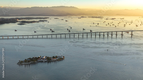 Aerial view of the Rio-Niteroi Bridge spanning Guanabara Bay with Ilha Fiscal, anchored ships, and mountains under a golden sunset in Rio de Janeiro, Rio de Janeiro, Brazil.