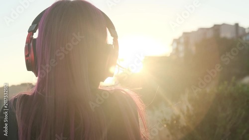 Rear view of a young woman wearing large wireless headphones and admiring the sunset over the city. In harmony with urban movement and music. Wind blowing hair, sun flare, relaxing melody, city vibe.